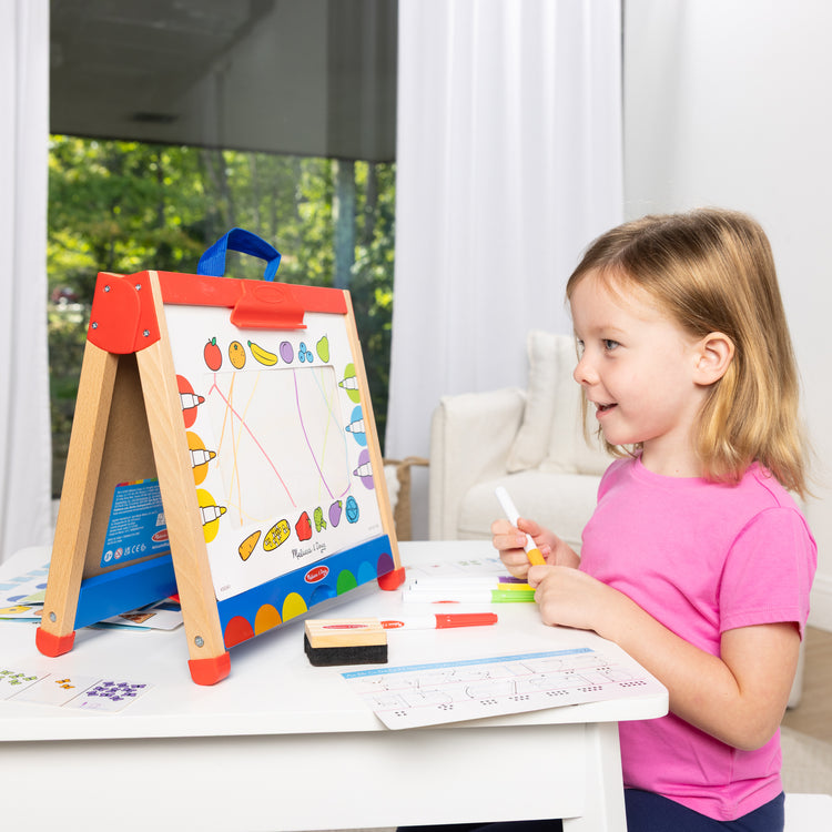 A kid playing with The Melissa & Doug Take-Along Double-Sided Wooden Tabletop Activity Art Easel; Portable Travel Toy with Whiteboard, Chalkboard, Arts and Crafts Accessories
