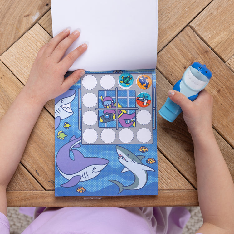 A kid playing with The Melissa & Doug Sticker WOW!™ Shark Bundle: 24-Page Activity Pad, Sticker Stamper, 500 Stickers, Mini Pad, Arts and Crafts Fidget Toy Collectible Character